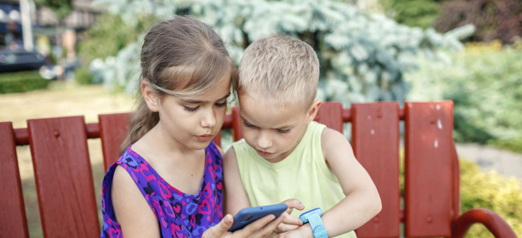 A young girl and a young boy sitting on a bench looking at a smartphone and a smartwatch.